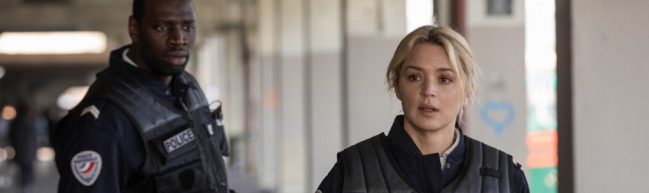 Aristide und Virginie in Polizeiuniform. Virginie läuft ein paar Schritte vor Aristide und er schaut ihr hinterher.