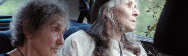 Zwei ältere Personen sitzen auf der Rückbank eines Autos. Die Person links hat kurze, graue gelockte Haare und blickt nach vorne. Die Person rechts hat lange, graubraune Haare, trägt ein geblümtes Nachthemd und blickt mit weit geöffneten Augen aus dem Autofenster. Sie hält einen Wiesenblumenstrauß.