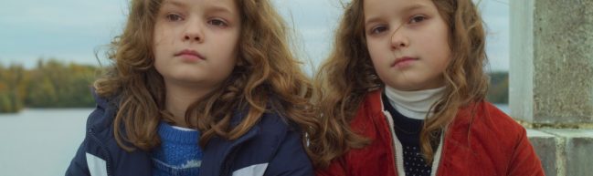 Nelly und Marion im Alter von 8 Jahren - zwei blonde Mädchen mit langen lockigen Haaren, die sich zum Verwechseln ähnlich sehen. Eine von ihnen trägt eine blaue, die andere eine rote Jacke.