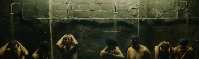 Eine Gemeinschaftsduschen im Gefängnis, karg und dunkel. Sieben Männer stehen in einer Reihe und duschen sich gleichzeitig.