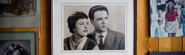 Eine Holzwand mit verschiedenen Fotografien. Im Zentrum eine gerahmte Fotografie in schwarz weiß, die eine Frau und einen Mann zeigt, Anfang 20 etwa, beide adrett gekleidet blicken sie seitlich aus dem Bild heraus.