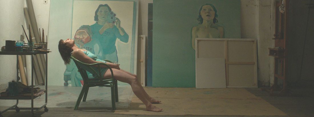 Minichmayr als Lassnig in ihrem Atelier. Sie sitzt auf einem Korbsessel, weit zurückgelehnt mit geschlossenen Augen. Sie trägt ein blaues Unterhemd. Neben ihr ein Regal mit Malutensilien. Im Hintergrund Gemälde in Blautönen.