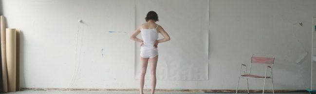 Maria Lassnig in ihrem Atelier. Sie steht mit dem Rücken zu uns, trägt ein Unterhemd und eine Unterhose, stemmt die Hände in die Hüften. Vor ihr an der weißen Wand hängt ein großes weißes Blatt Papier.