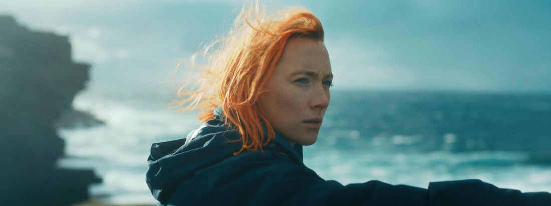 Portraitaufnahme der Hauptfigur Nora, gespielt von Saoirse Ronan, mit vom Wind zerzaustem Haar und entschiedenem, nach rechts gewendetem Blick. Im Hintergrund unscharf Klippen und das Meer.