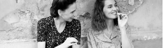 Zwei Frauen, eine davon Delia, vor einer beschädigten Mauer. Sie tragen geblümte Blusen und rauchen Zigaretten. Sie lächeln.